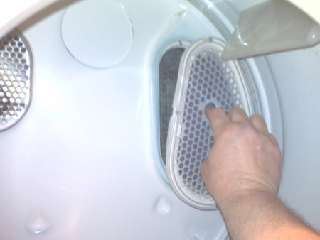 The interior of a Whirlpool dryer is shown. A hand is reaching in, with the pointer finger and thumb inserted in the holes for the lint trap, which is loosened from its housing but not fully extricated.