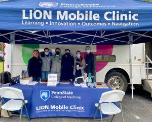 Tent and table with LION Mobile Clinic and Penn State College of Medicine logos, with students standing behind the table and a bus behind them.